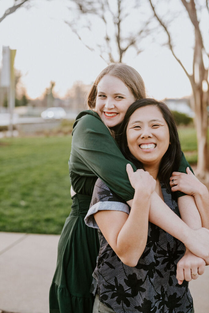 A person standing behind their partner with their arms around them as they both smile 