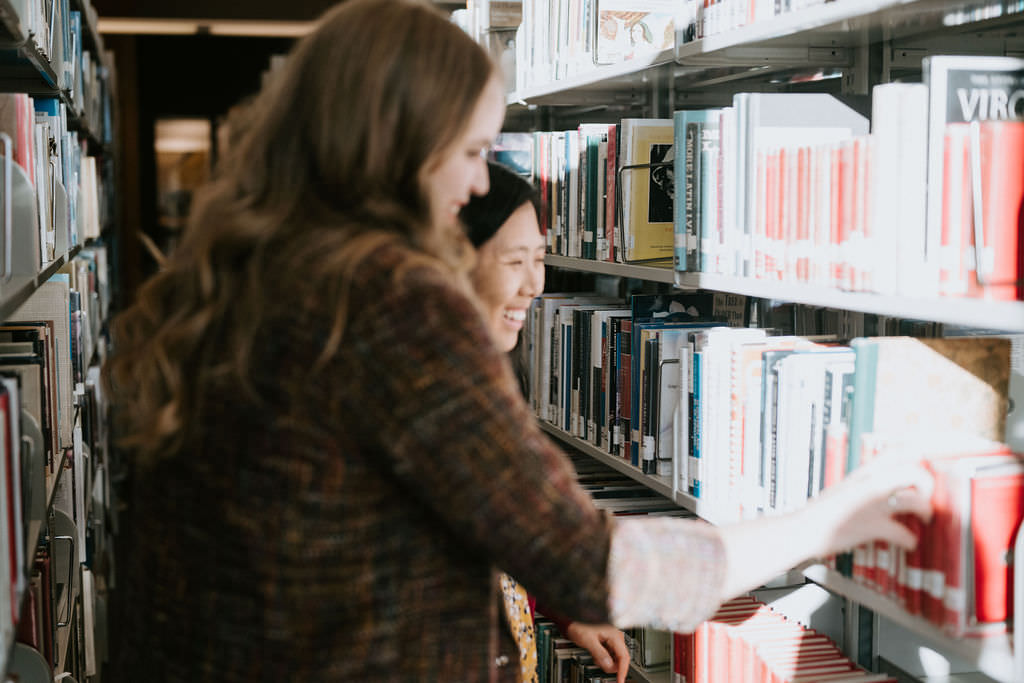 A couple during their library engagement photos standing next to a shelf of books looking at them and smiling