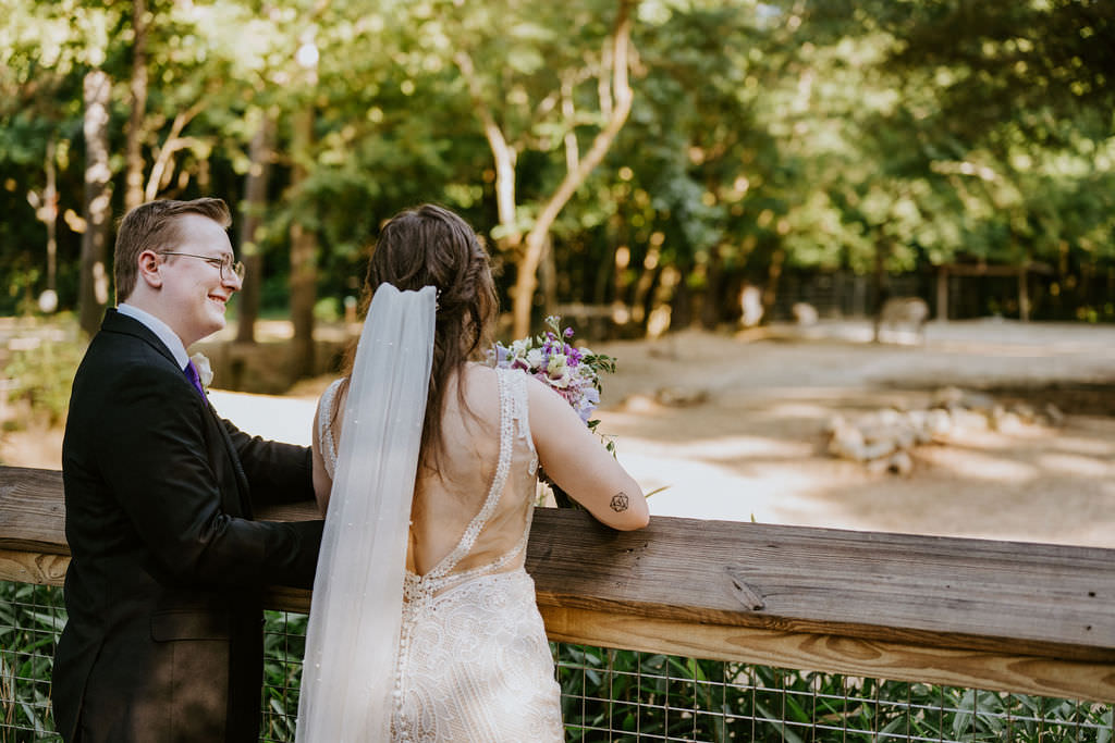 A newlywed couple standing by a wooden railing, smiling and looking at zoo animals in the distance.