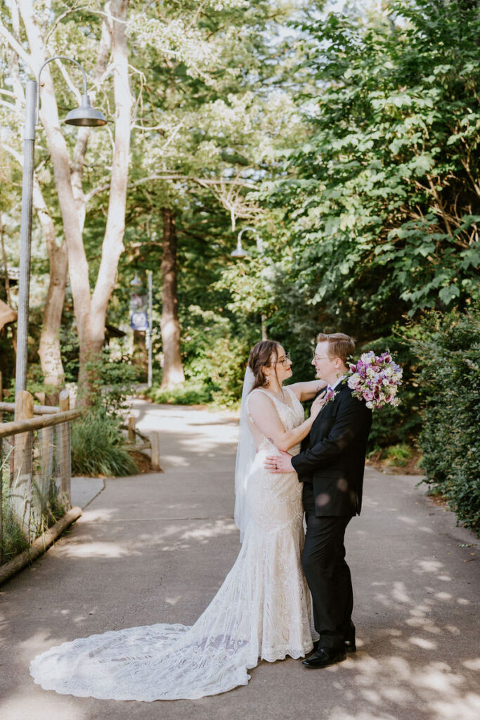 A wedding couple with their arms around each other on a shaded pathway surrounded by trees.
