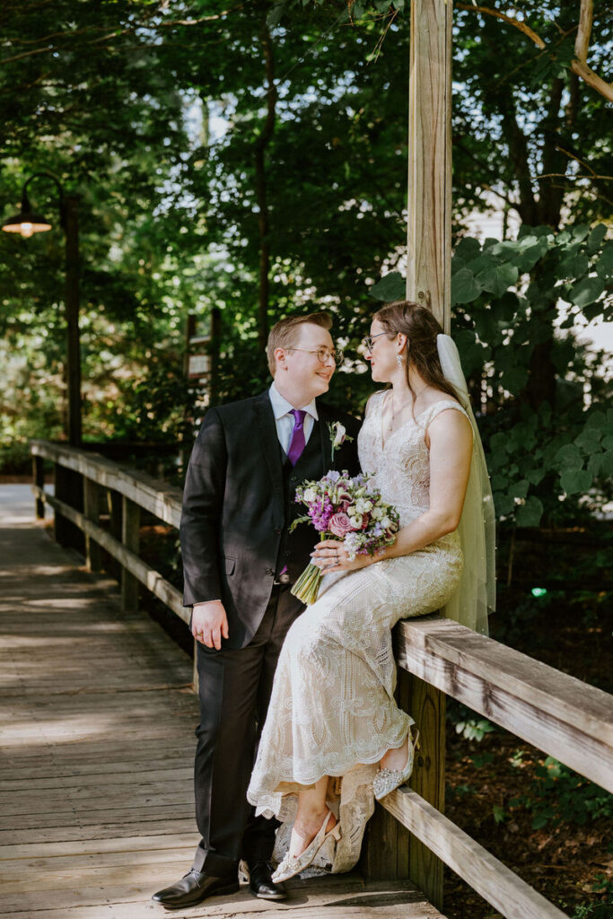 A newlywed couple sitting on a wooden bridge, gazing into each other's eyes lovingly.