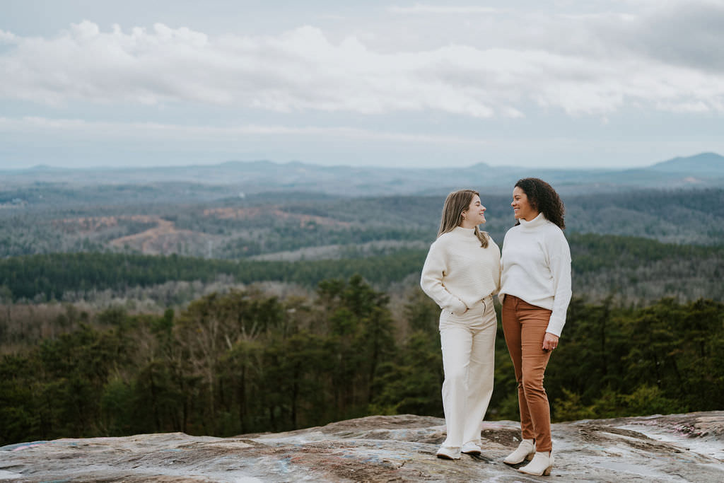 Two women stand side by side on a mountain overlook, smiling at each other with the forested landscape behind them