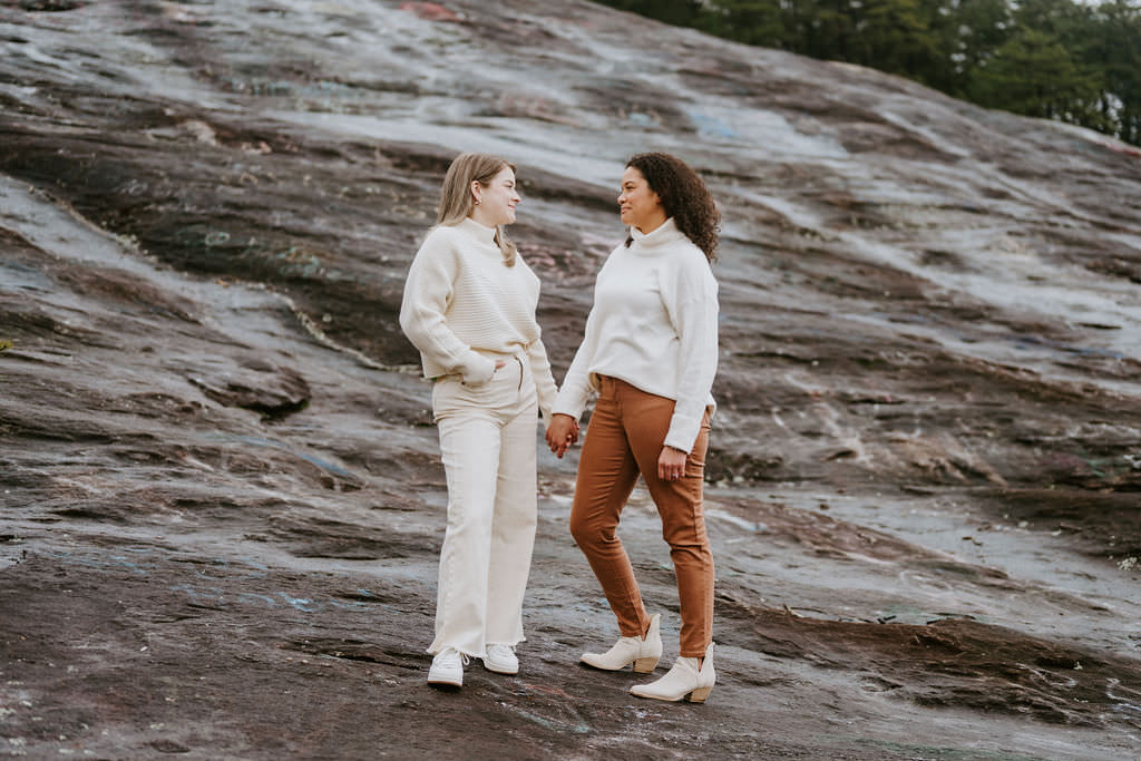 Two women standing on rocky terrain, facing each other while holding hands.
