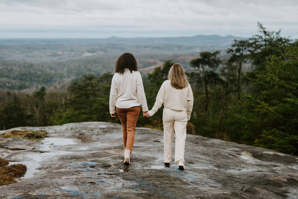 A couple holding hands while standing on a rocky mountain surface with a panoramic view.
