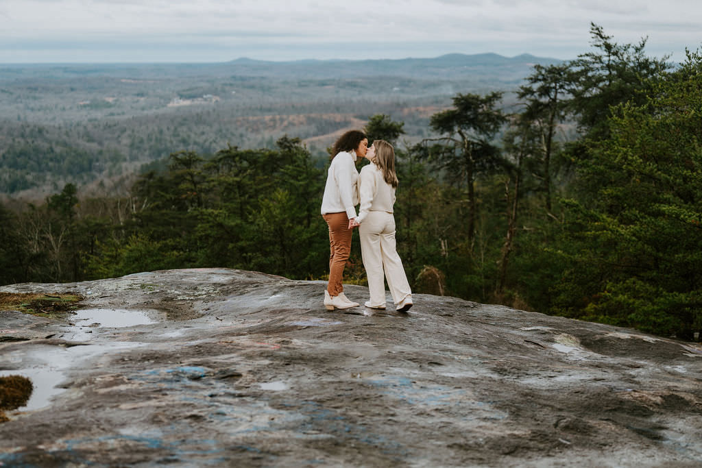 A couple standing on a mountain overlook, kissing while holding each other close.
