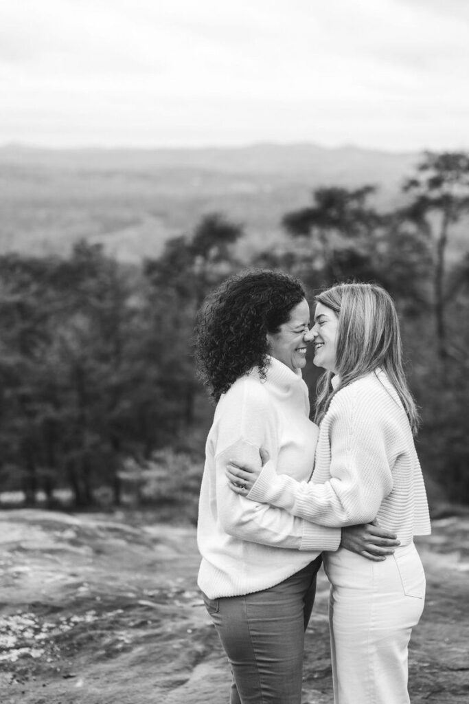 A black and white photo of two women smiling and embracing with a forested mountain view in the background.
