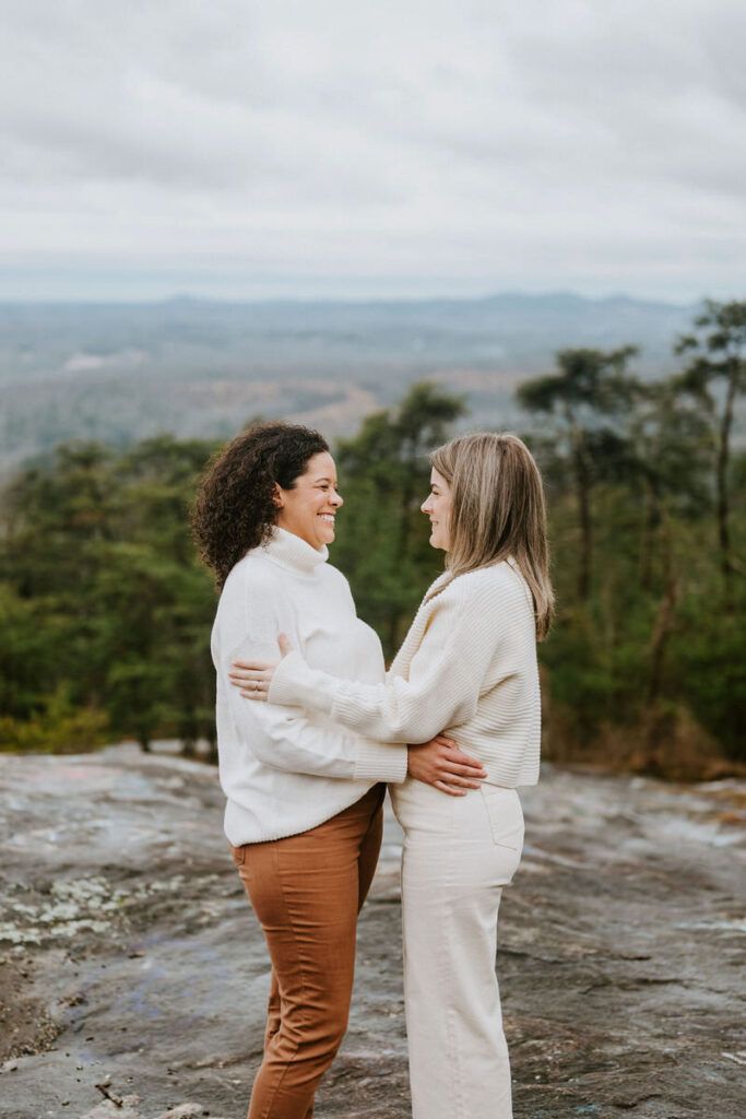 A couple stands facing each other, smiling, with their arms around each other on a mountain overlook.
