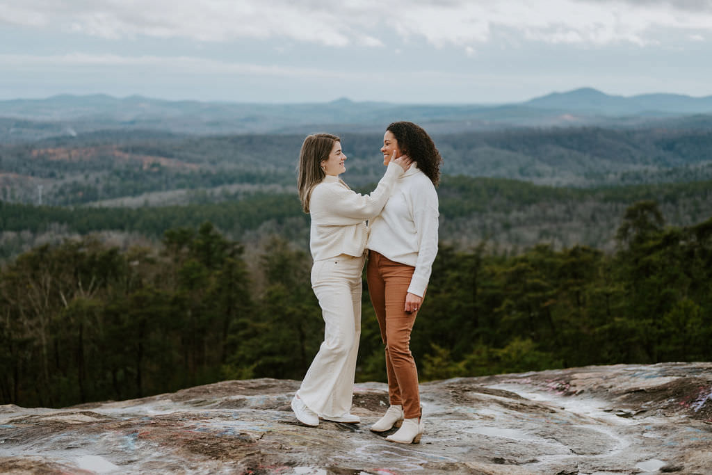 One woman tenderly touches the other's cheek as they stand on a rocky mountain surface