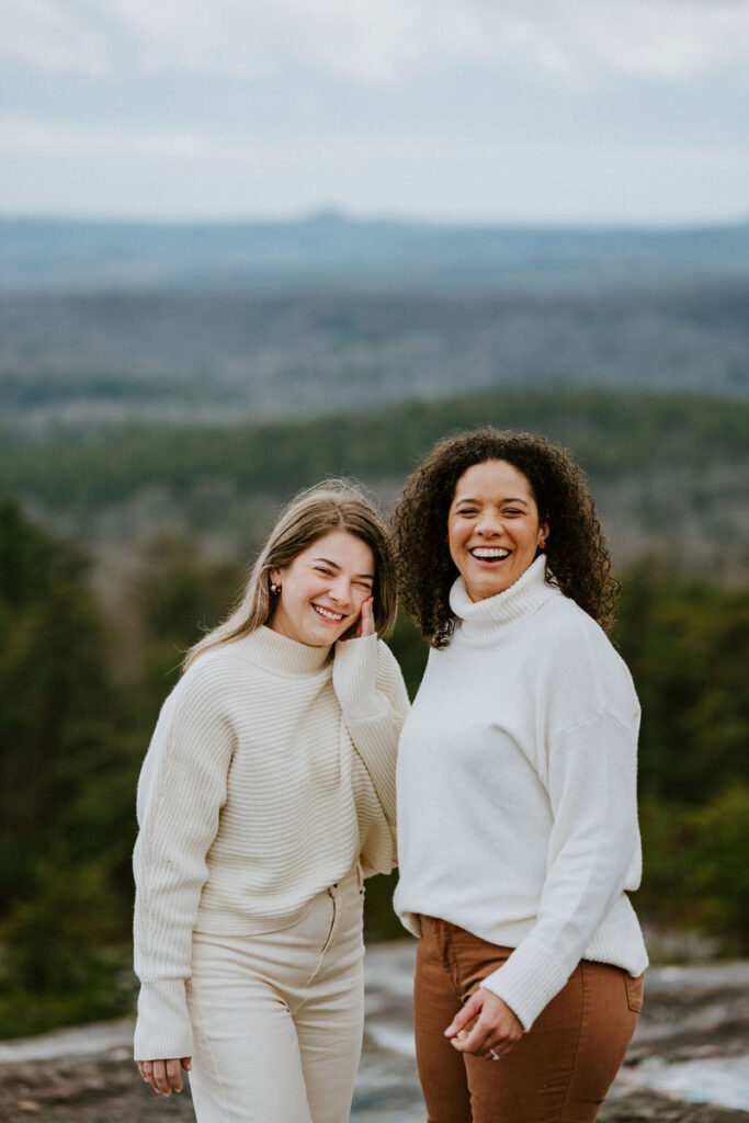 Two women laughing and smiling, standing close together on a mountain overlook.
