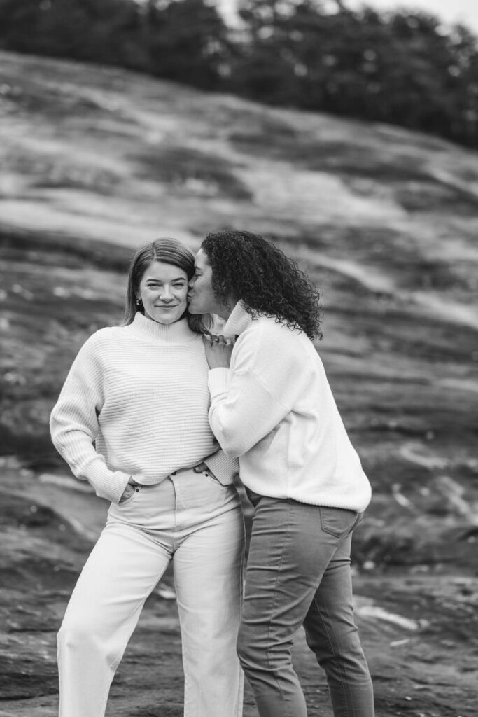 One woman kisses the other's cheek while they stand on rocky terrain