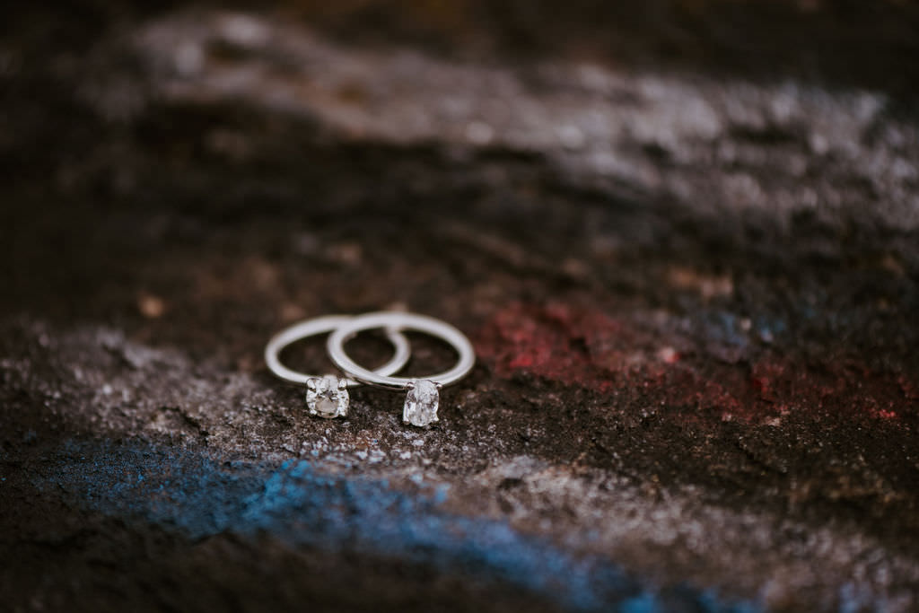 A close-up of two engagement rings resting on a textured rock surface with hints of blue and red colors.
