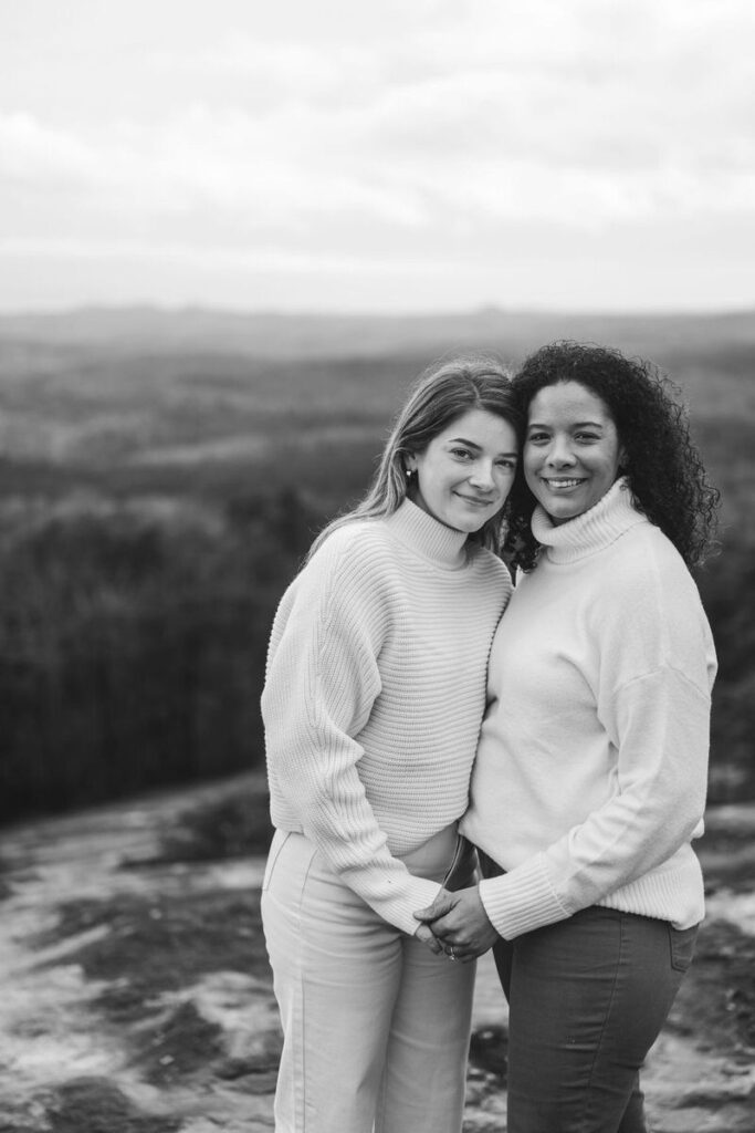 Two women stand close together, holding hands and smiling, with a mountainous landscape in the background