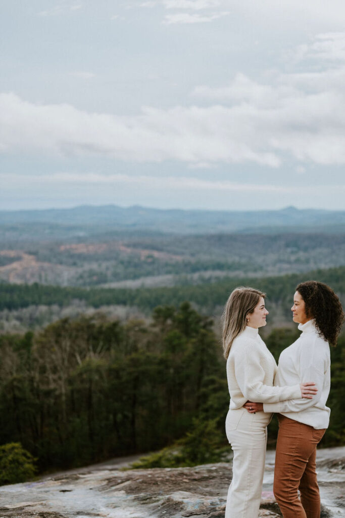 A couple stands on a mountain overlook, facing each other and smiling