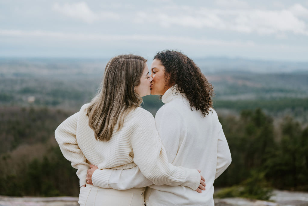 Two women in sweaters kiss during mountain engagement photos.