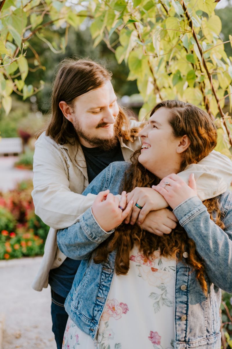 A man has his arms wrapped around a woman's shoulders and they are both looking at each other and smiling the woman has on a blue jean jacket and a white dress with a floral pattern and the man has on a blue t-shirt and a tan button-up 