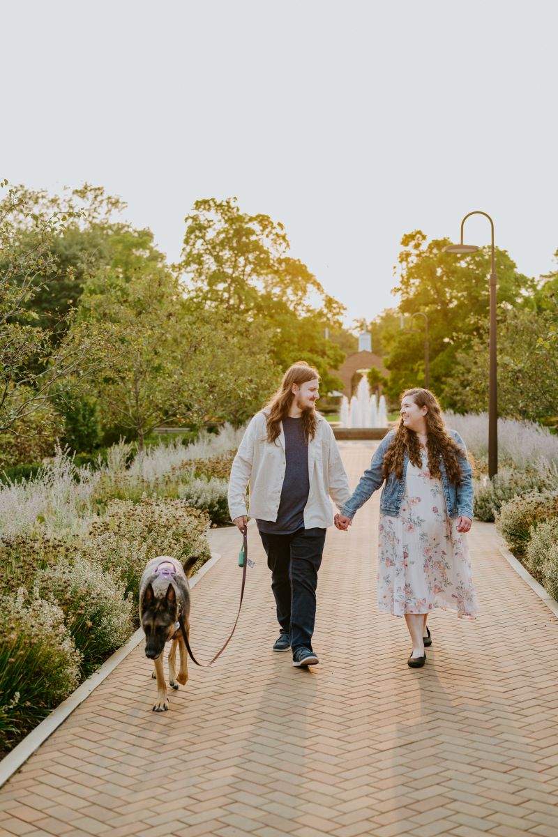 A man and woman are walking along a paved path with their dog and holding hands and looking at each other behind them is fountain and trees and shrubs 