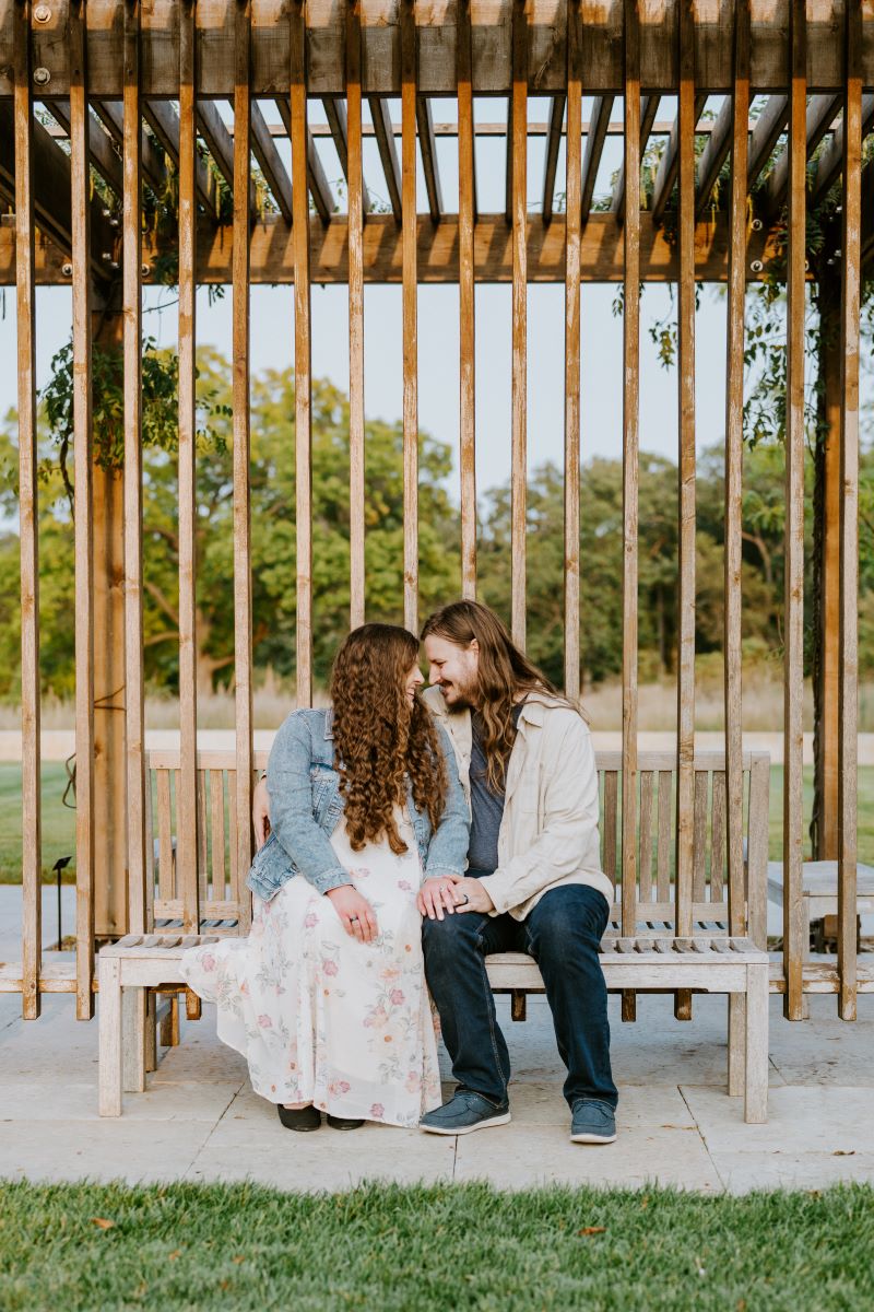 A man and woman are sitting on a wooden bench and have their heads touching each other and they are both smiling and holding hands the woman has on a white dress with a floral pattern and a jean jacket and the man is wearing a tan button up and jeans