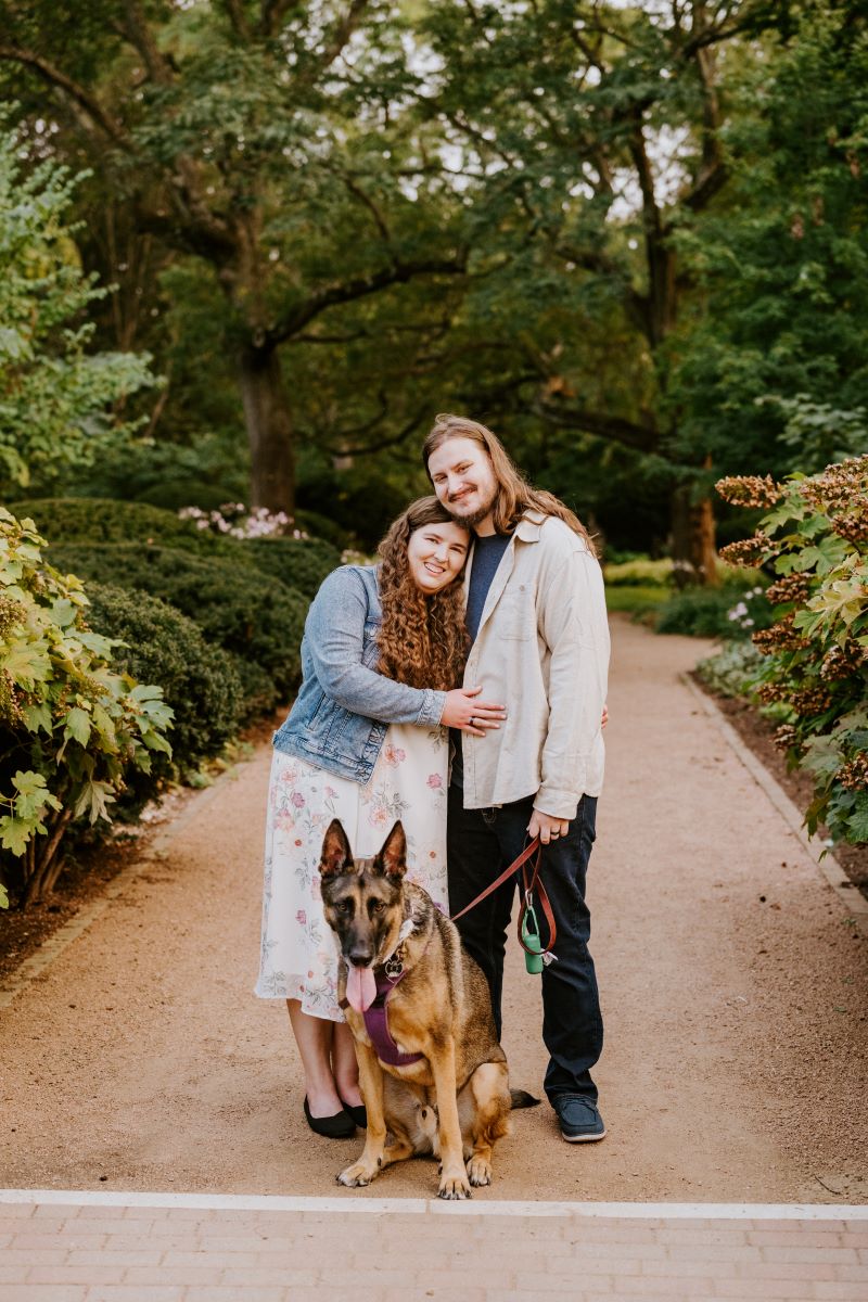 A man wearing a tan button up, a blue shirt, and jeans is holding the red leash of his dog and his partner is giving him a side hug and both are smiling and she is wearing a blue jean jacket and a white dress with a floral pattern