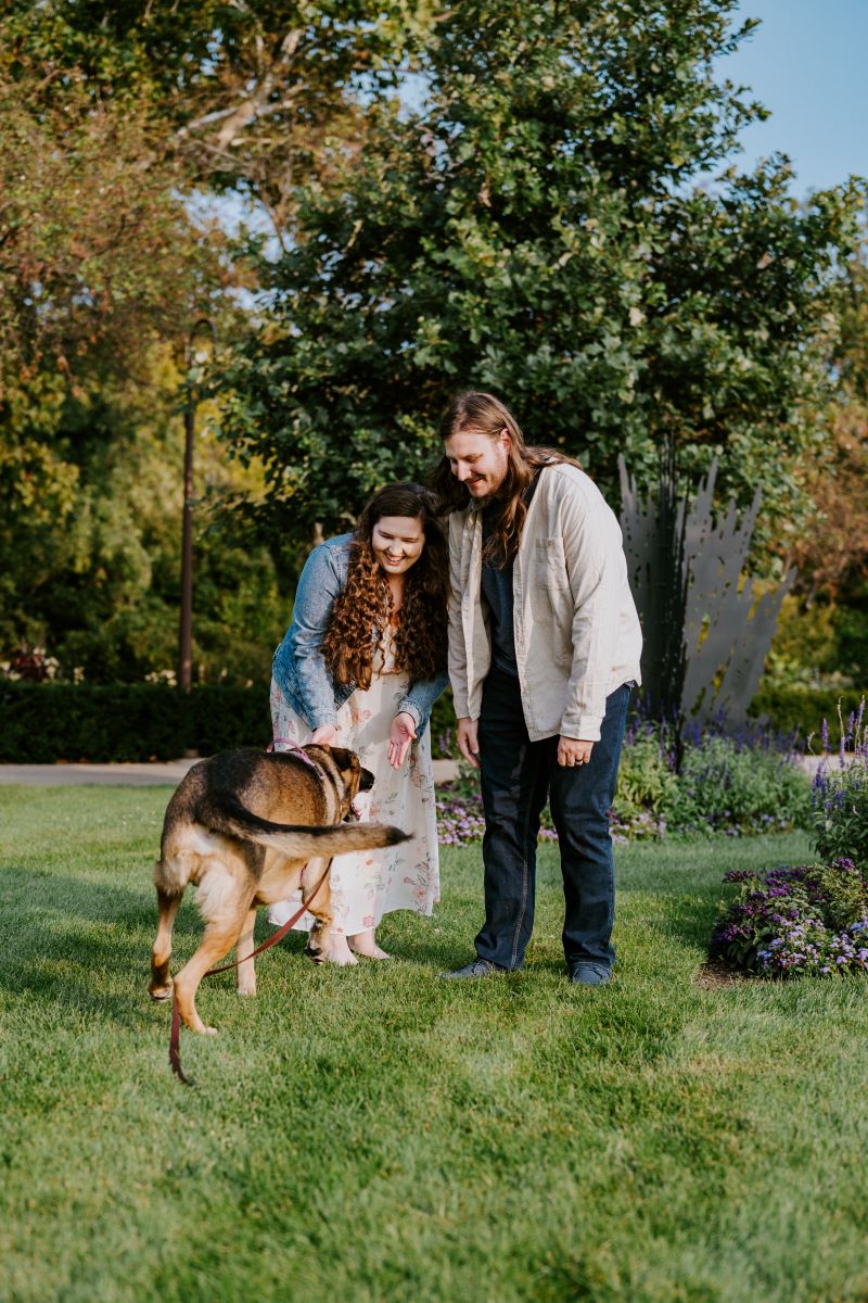 A man wearing a tan button up, blue jeans, and a blue shirt is leaning over and looking at his dog who is running towards him with his partner who is wearing a white dress with a floral pattern and a jean jacket and smiling as the dog comes running towards them