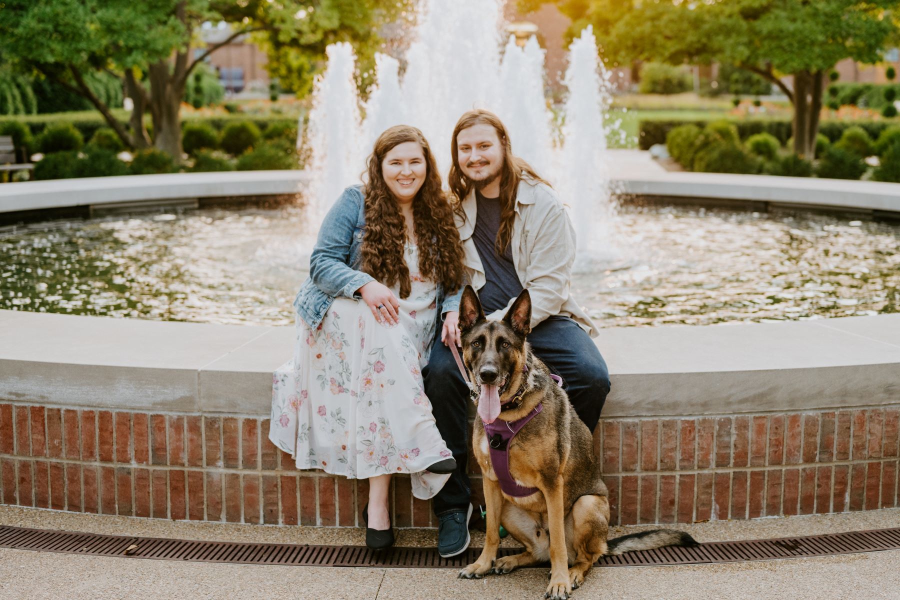 A man and a woman are sitting on the edge of a fountain with their dog the man and woman are both smiling and the woman is wearing a white dress with a floral pattern and a jean jacket and the man is wearing a blue shirt and a tan button up and blue jeans