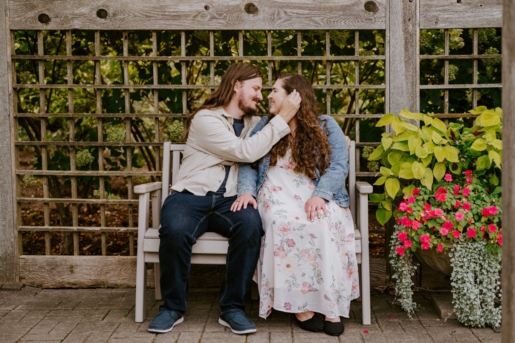 A man and a woman are sitting on a bench the man is tucking the woman's hair behind her ear and smiling and the woman is touching his thigh the woman is wearing a floral dress and a jean jacket and the man is wearing a tan button up, a blue t-shirt, and blue jeans and they are surroudned by potted plants and flowers 