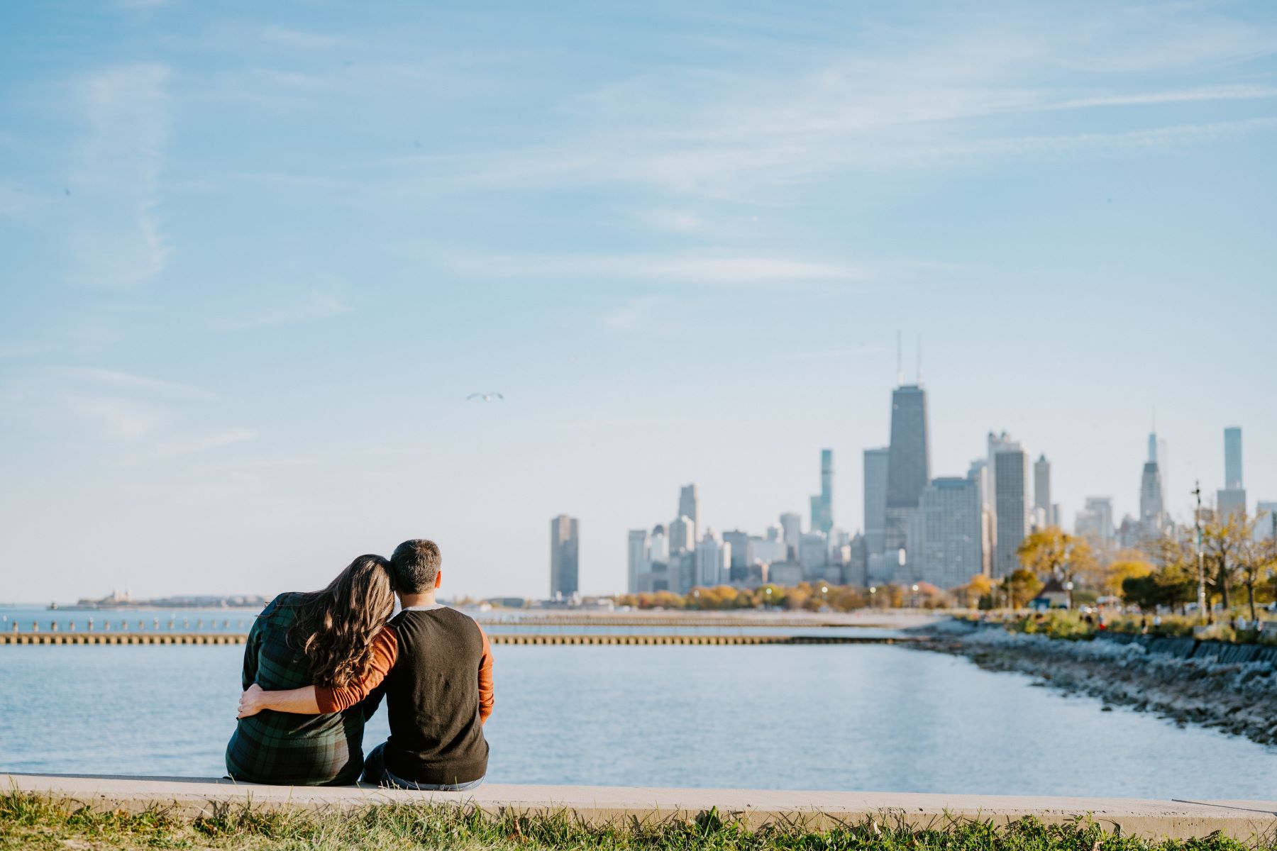 A couple sitting together in front of a lake looking at a city skyline the woman has her head on her partner's shoulder and her partner has his arm wrapped around her 