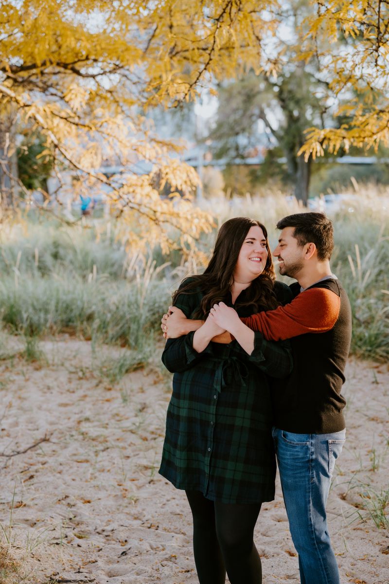 A couple standing underneath a tree that has yellow leaves and the are smiling at each other the woman is wearing a green and black flannel dress and black tights the man is wearing a black and red shirt and blue jeans and he is hugging his partner and she is holding his arm