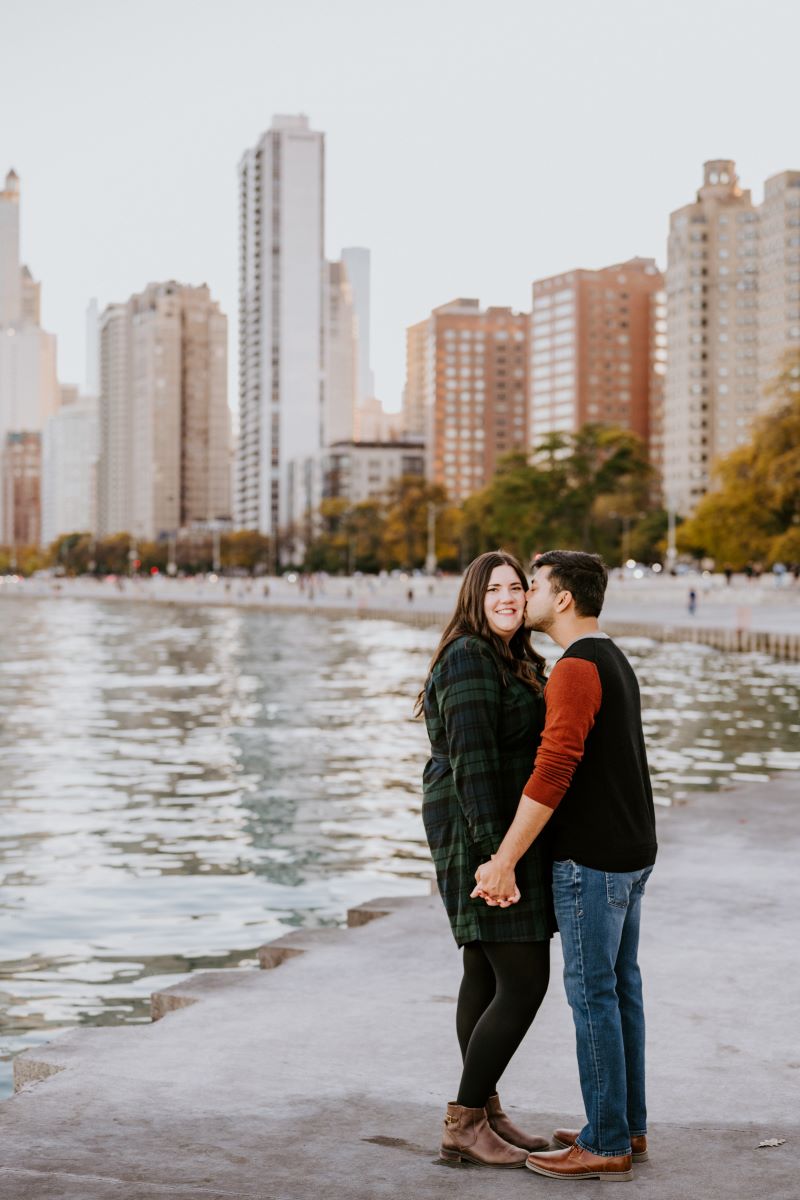 A copule standing next to a lake with a city skyline behind them and the woman is wearing a green and black flannel dress and black tights and smiling and the man is kissing her cheek wearing a black and red shirt an dblue jeans 