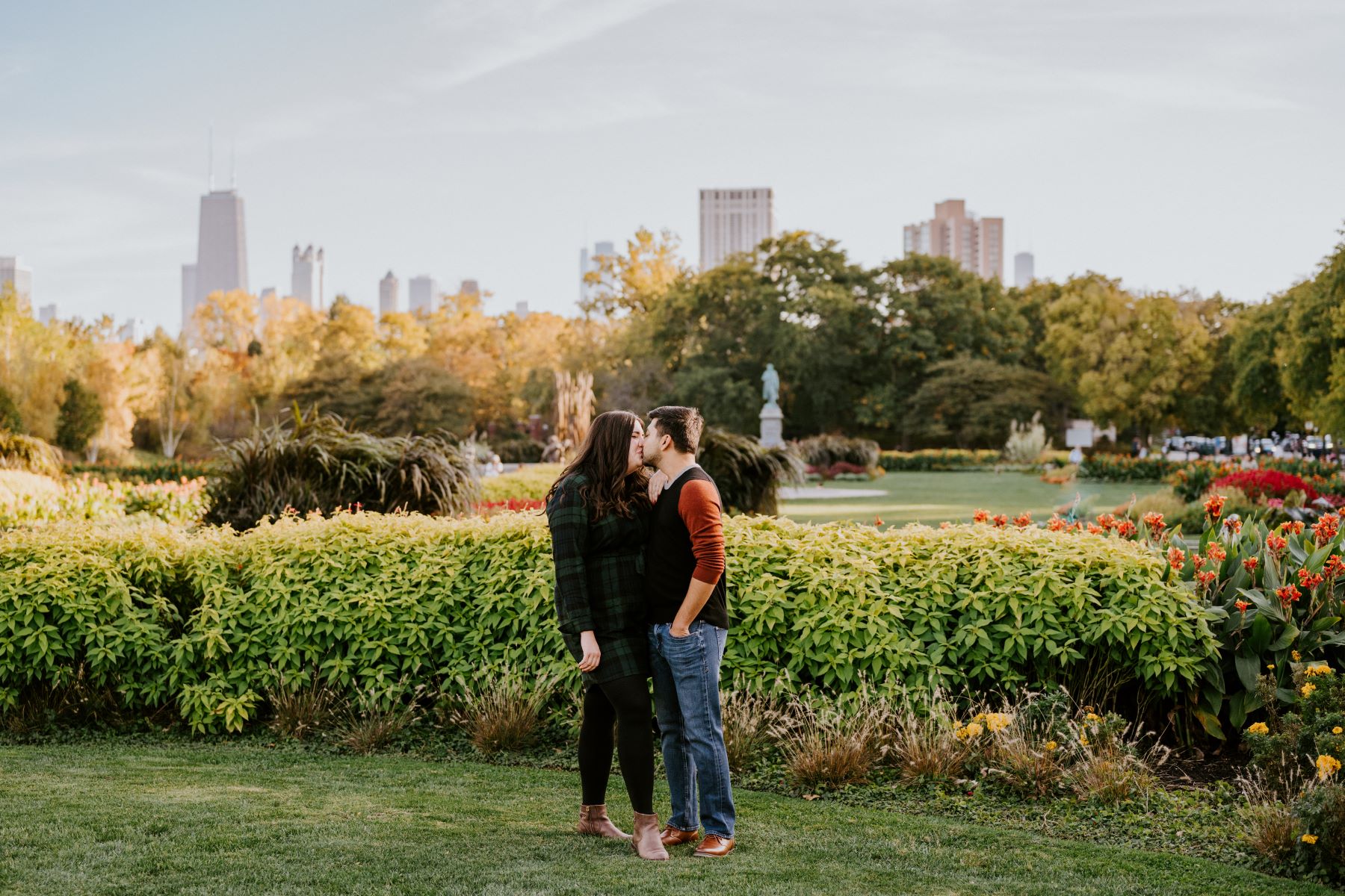 A couple kissing in a park the woman is wearing a green flannel dress the man is wearing a red and black shirt and blue jeans and behind them are plants and a city skyline