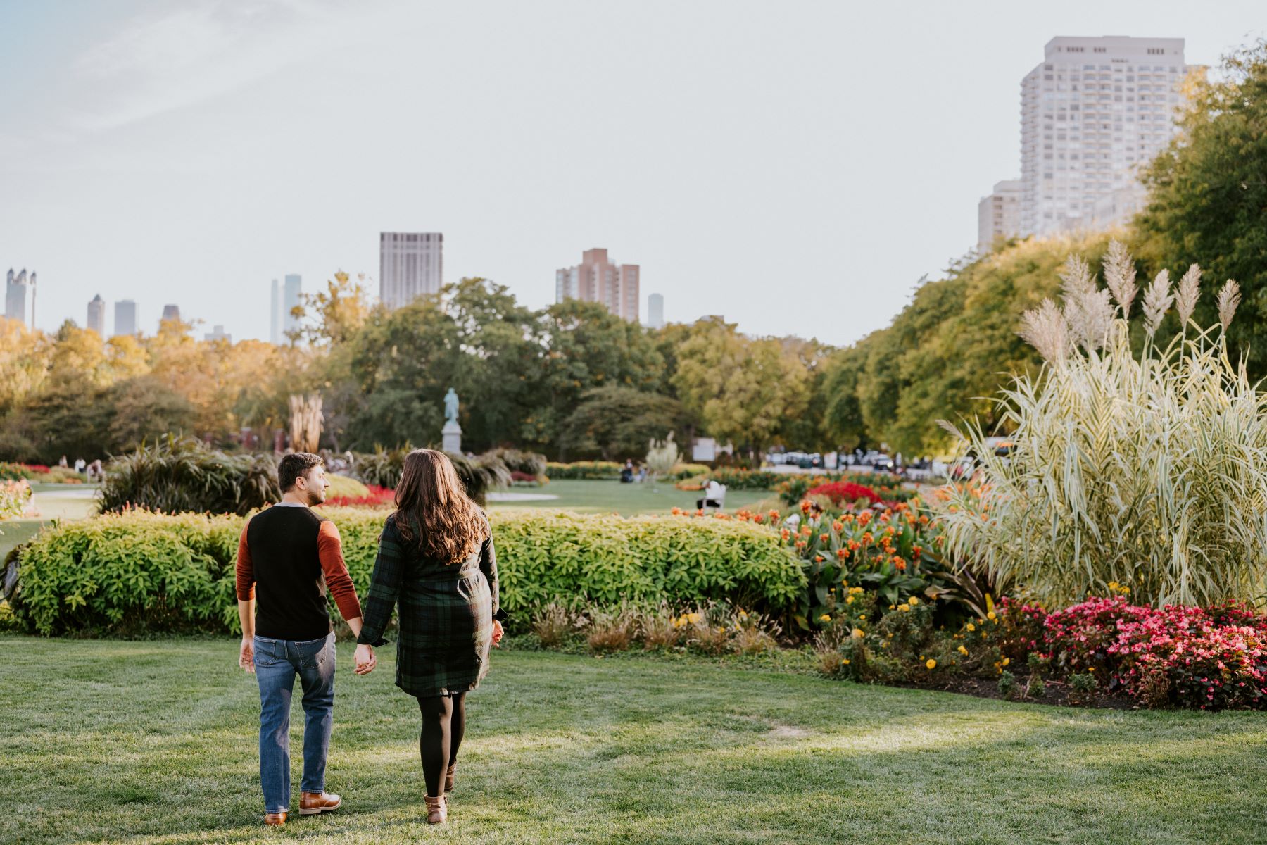 A couple walking together in a park looking at each other and holding hands the woman is wearing a green flannel dress and the man is wearing a red and black shirt and jeans and in front of them are plants and a city skyline