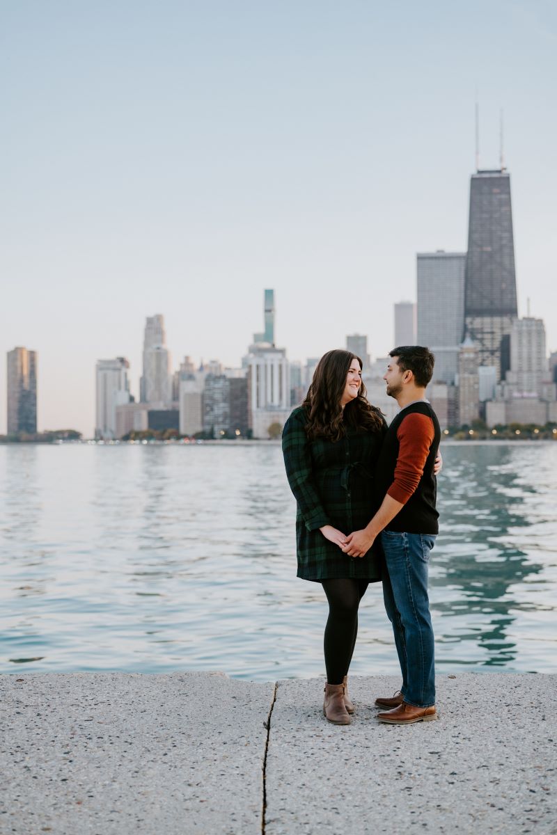 A couple smiling at each other and standing next to a lake with a city skyline behind them they are also holding hands and the woman is wearing a black and green dress and black tights and the man is wearing a red and black shirt with blue jeans