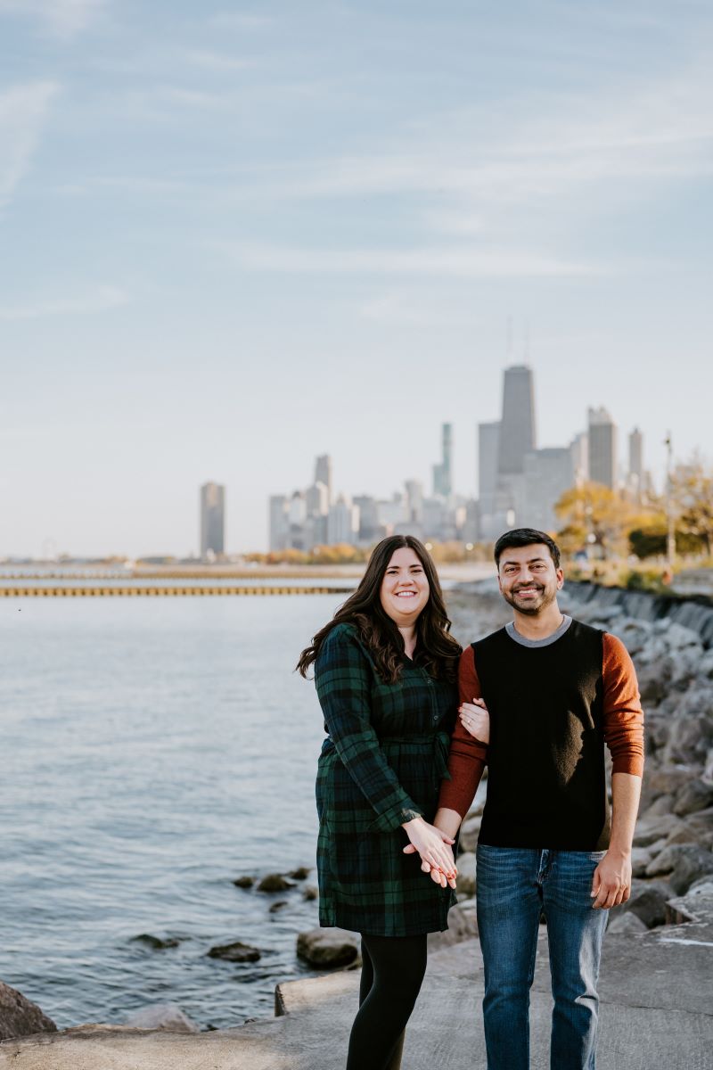 A couple smiling and standing next to a lake with a city skyline behind them the woman is wearing a black and green flannel dress and the man is wearing a red and black shirt with blue jeans