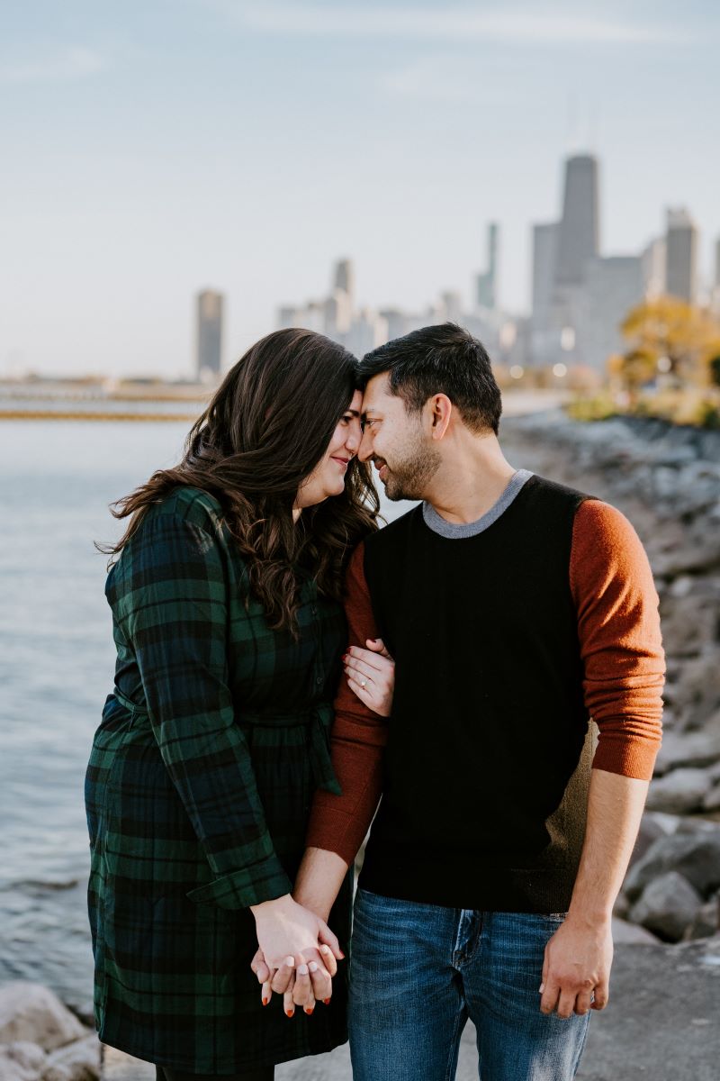 A couple pressing their foreheads together and smiling the woman is wearing a green and black flannel dress and the man is wearing a red and black shirt and jeans and they are holding hands behind them is a lake and a city skyline