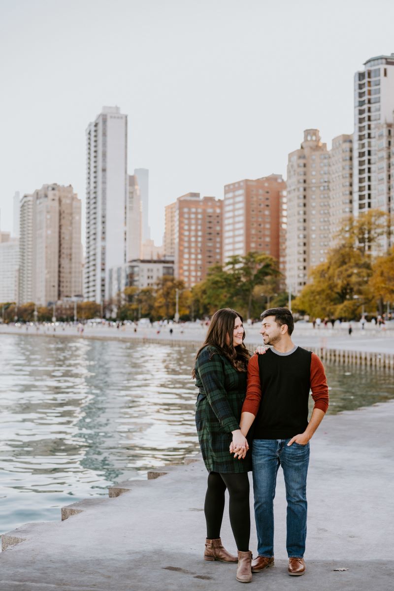 A couple standing next to a lake with a city skyline behind them they are holding hands and the man is wearing a black and red shirt and blue jeans the woman is wearing a green and black flannel dress 