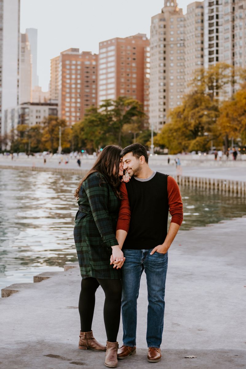 A couple standing next to a lake laughing together they are holding hands and the woman is leaning towards her partner the woman is wearing a green and black flannel dress and black tights and the man is wearing a red and black shirt and blue jeans 