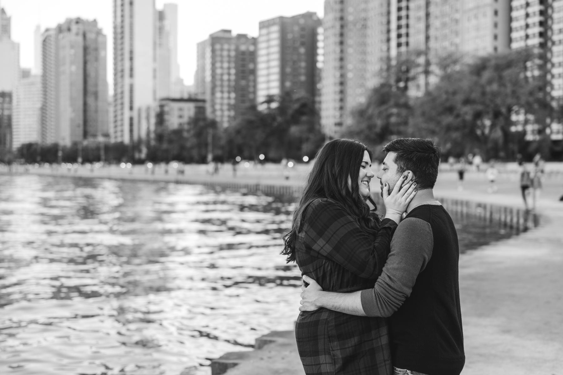 A couple standing next to a lake hugging each other with a city skyling behind them the man has his arms wrapped around the woman and the woman is holding the man's face with one hand