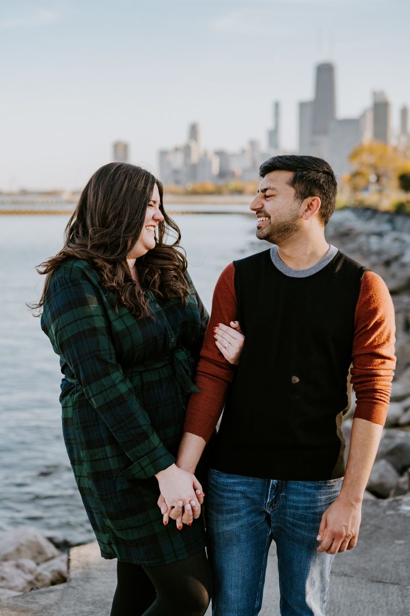 A couple laughing with each other the woman is holding her partner's arm and hand and is wearing a black and green flannel dress and the man is wearing a black and red shirt and blue jeans and behind them is a lake and city skyline