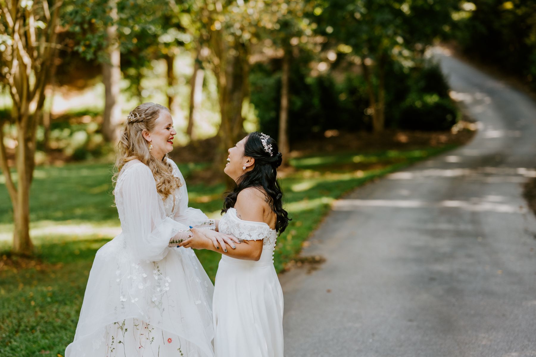 Two women wearing white wedding dresses laughing and holding each other's forearms and behind them is a paved road and trees