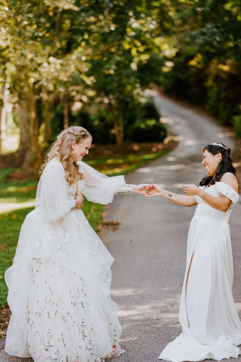 Two women in white wedding dresses on a paved road laughing with each other and holding hands 