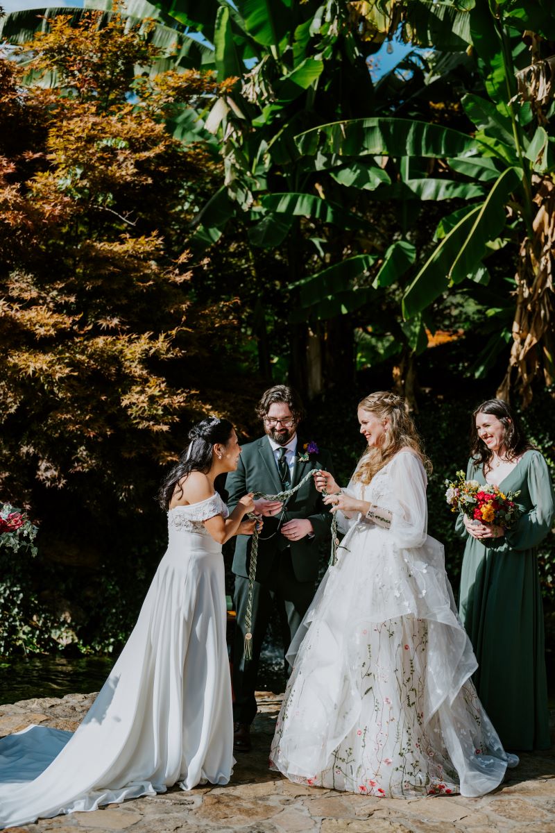 Two women in white wedding dresses at their wedding ceremony perfoming the handfasting tradition while holding a cord between them with their officiant standing behind them 