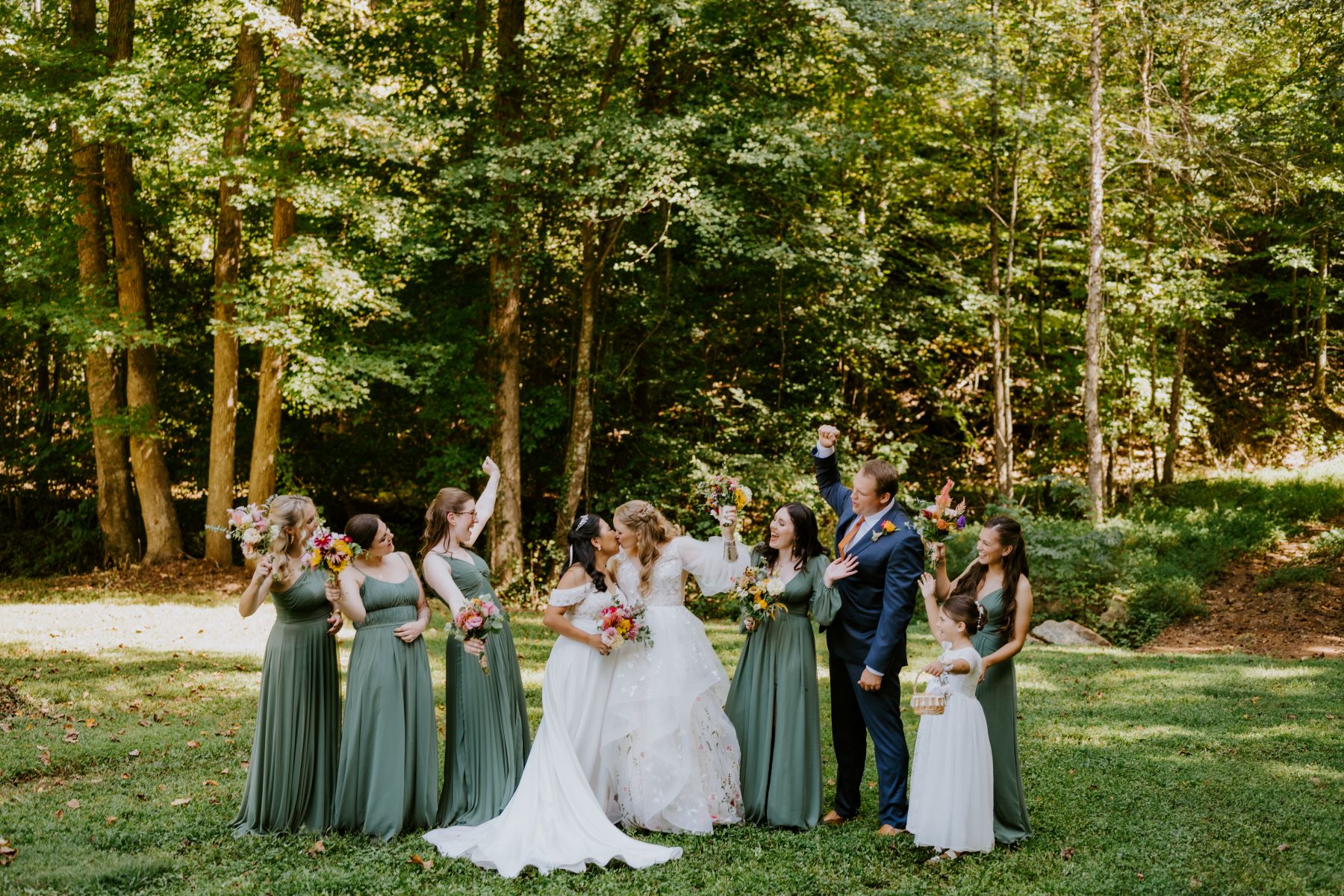 Two women in white wedding dresses kissing and holding bouquets of flowers while their guests are cheering for them while standing next to them