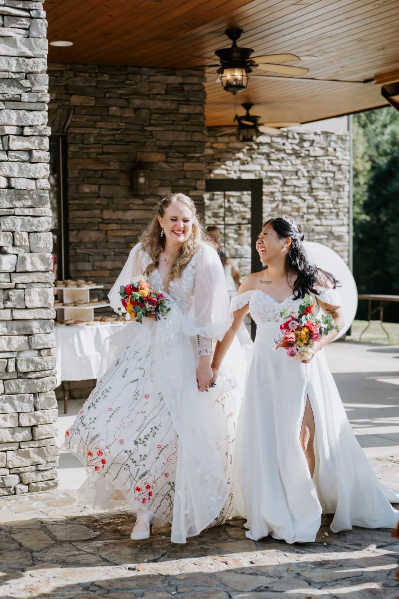 Two women wearing white wedding dresses holding hands and laughing and holding bouquets of flowers 