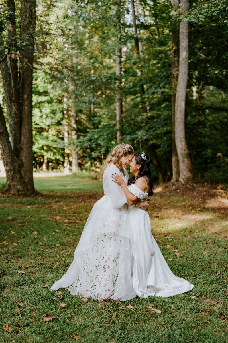 Two women wearing white wedding dresses with their heads pressed against each other smiling and looking into each other's eyes and they are surrounded by trees in a forest setting