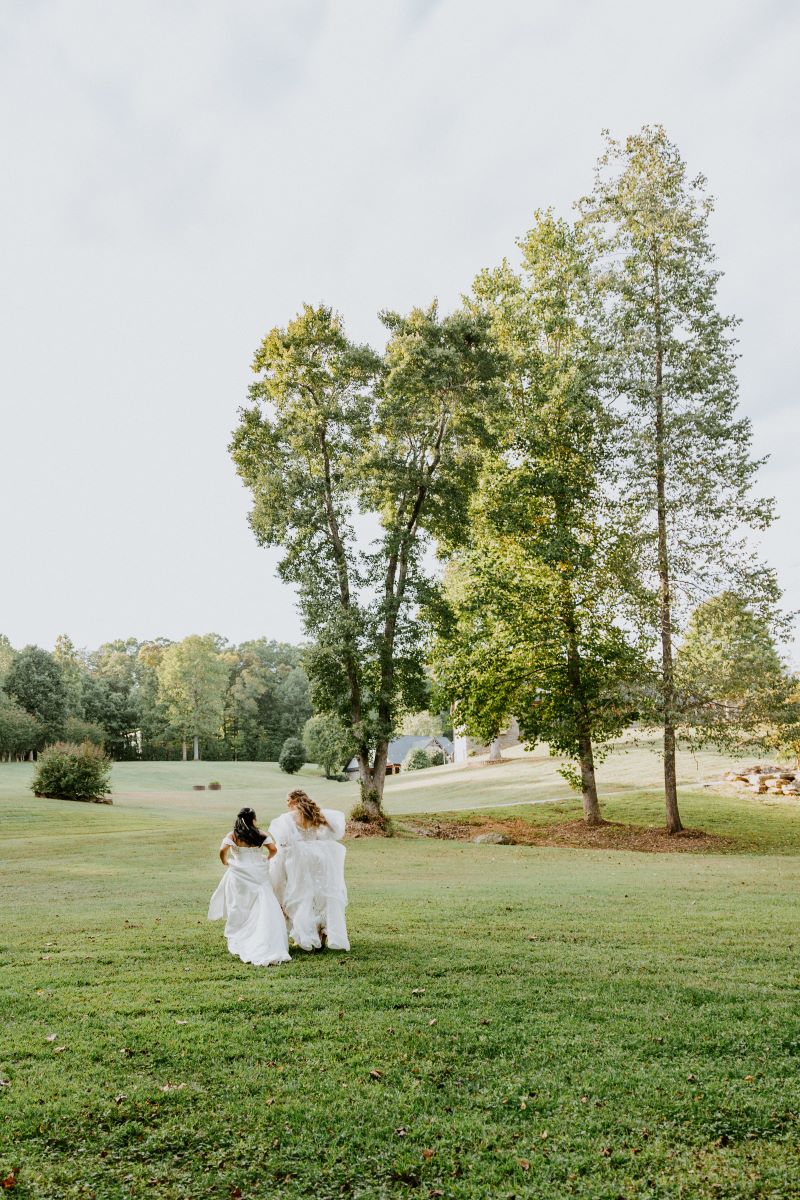 Two women in white wedding dresses walking in a green field of grass