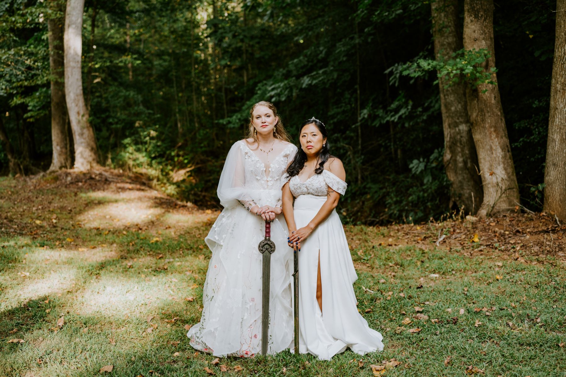 Two women in white wedding dresses in a forest both have swords they are holding in their hands 