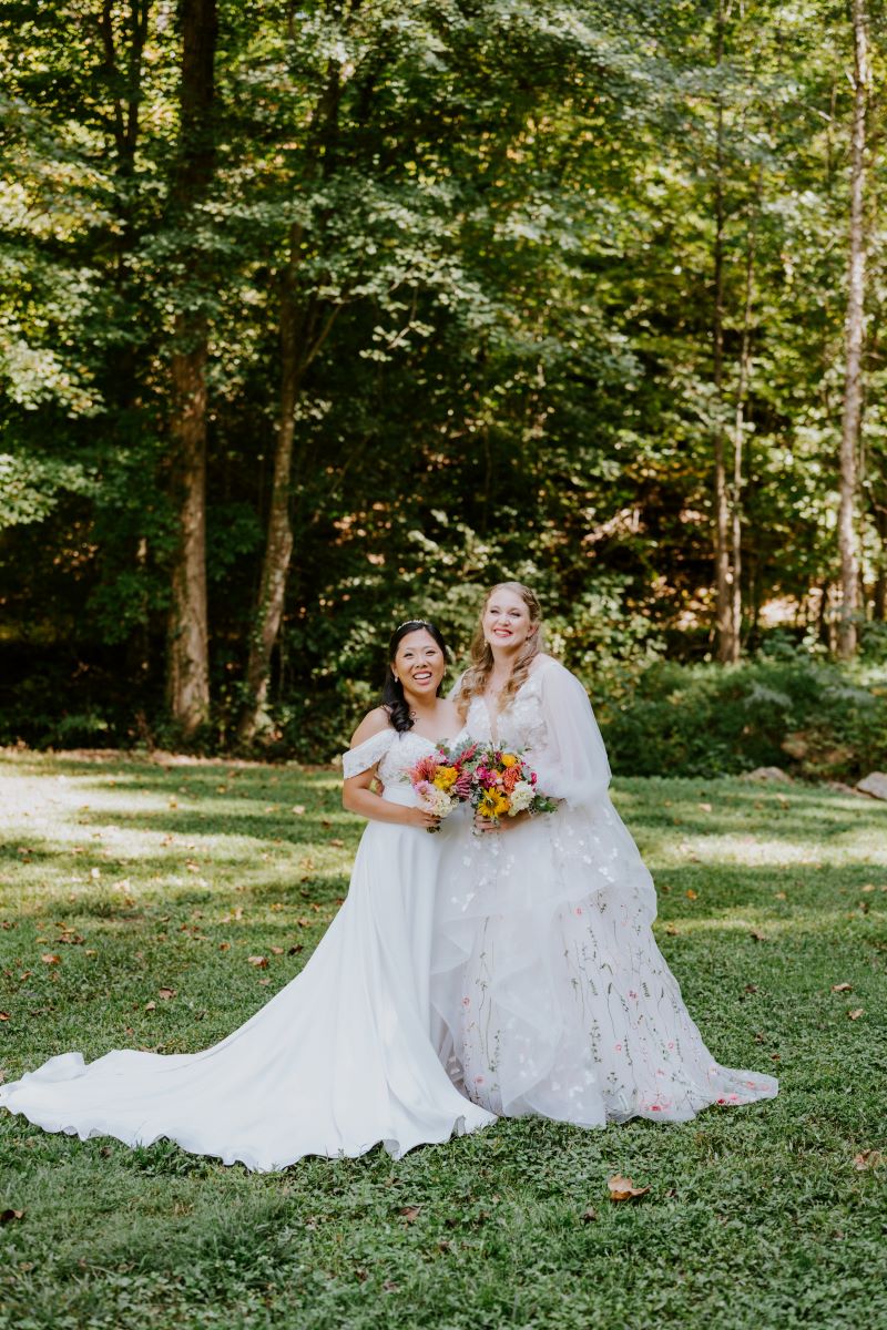 Two women in white wedding dresses in a forest hodling bouquets of flowers