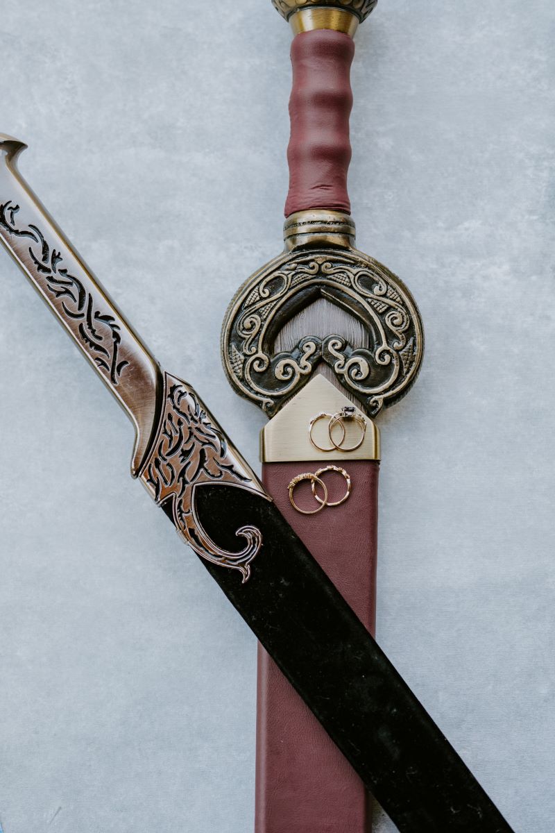 Two swords one is red and the other is black with gold handles with wedding rings laid on top of the red sword 