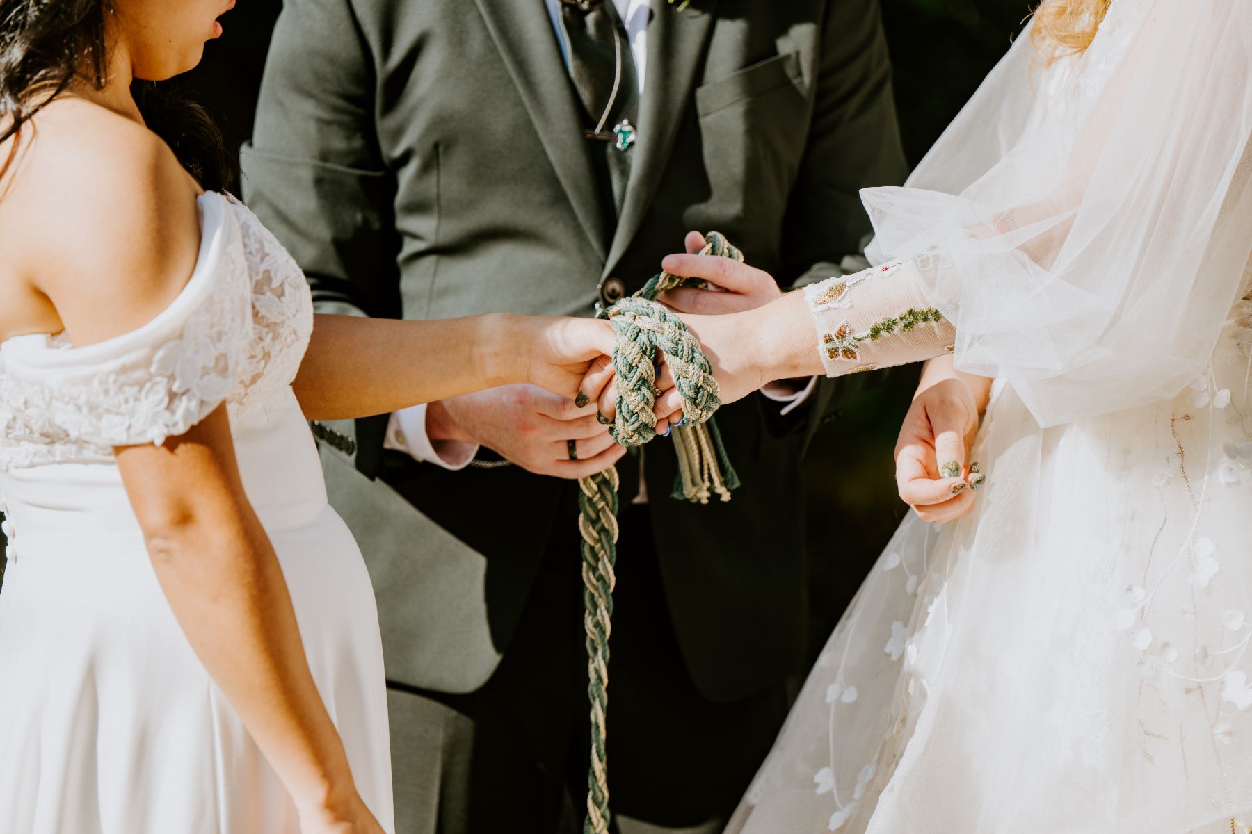 Two women in white wedding dresses holding hands with their wedding officiant behind them thee officiant is perfoming a handfasting tradition and wrapping a cord around both of their hands 