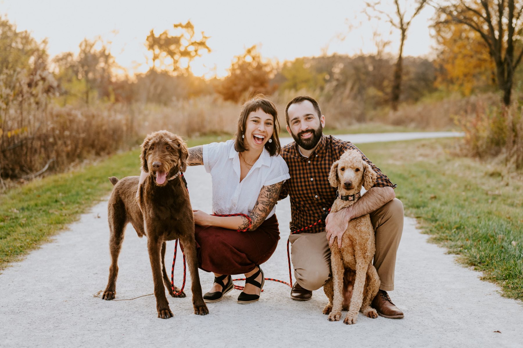 A woman wearing a white shirt and red pants is kneeling down next to her brown dog and her partner is wearing a plaid button up and tan pants and has his arm around her and with his other arm is holding their other brown dog the couples is kneeling down on a paved path 