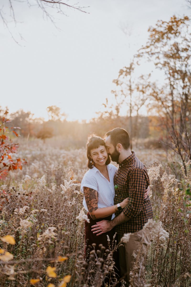 A couple hugging each other in a field the man has his head pressed against the woman's cheek and the woman is smiling 